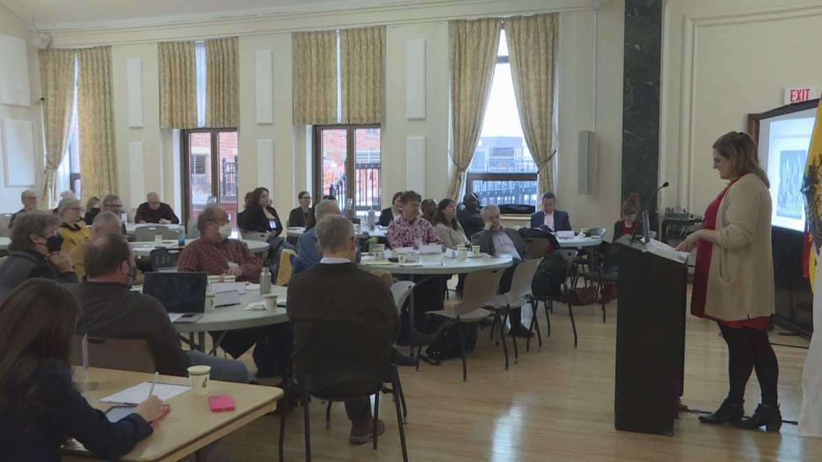 People sit at circular tables in a wide room, looking at a woman standing at the front of the room and speaking from a podium. 