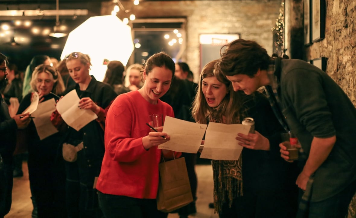 A group of three people crowd around two pieces of paper held by two of them, reacting with amusement at what they are reading. In the background, more people carrying more pieces of paper are visible. They are inside a building with rough brick and warm lighting.  