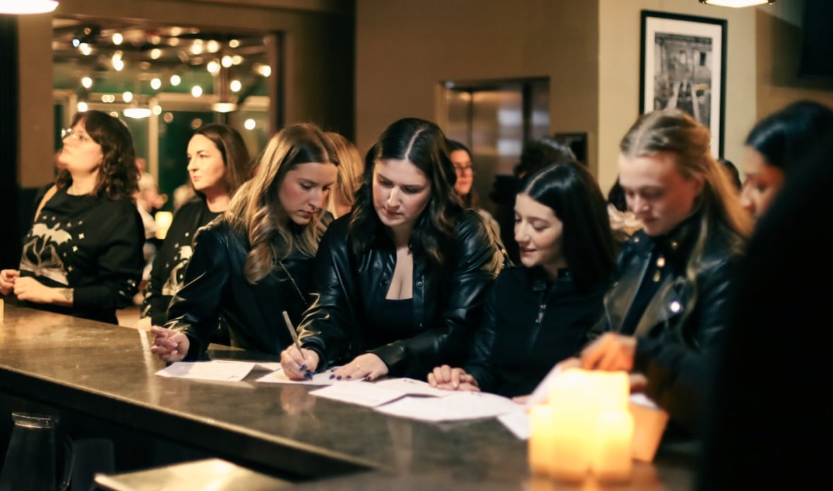 A group of woman, all wearing black, lean over pieces of paper sitting on a bar. A crowd is visible beyond them, and there is warm lighting throughout.  