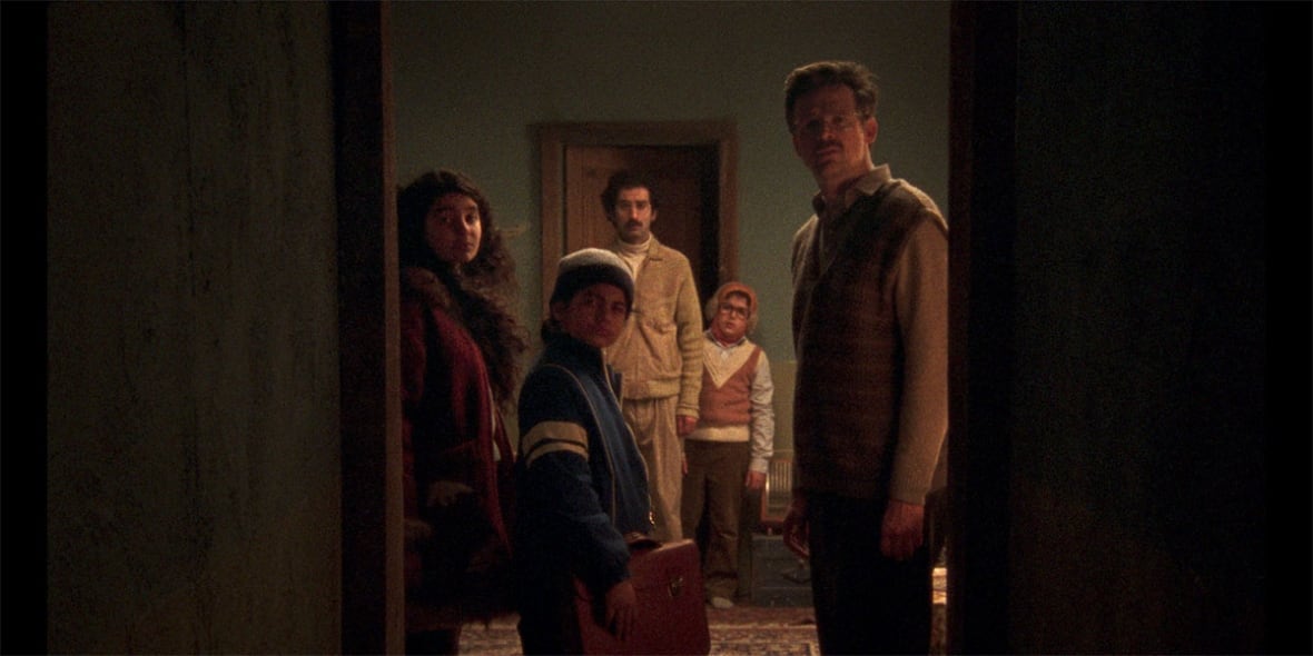 A still from a movie shows five people, two adults and three children, looking through a doorway.