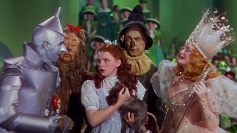 Judy Garland's Dorothy holding Toto and looking concerned in the middle of the Tin Man (Jack Haley), Cowardly Lion (Bert Lahr), Scarecrow (Ray Bolger), and Glinda the Good Witch (Billie Burke) in The Wizard of Oz