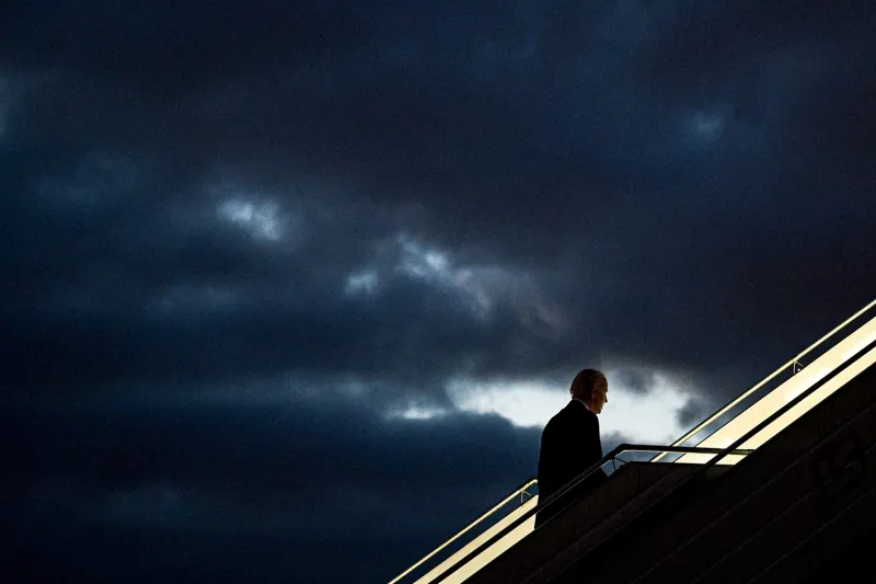 Joe Biden, in silhouette, walks up stairs to board a plane, with ominous dark clouds in the background.