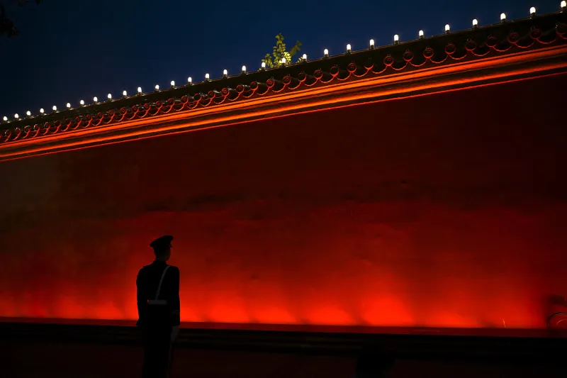 A silhouetted figure with a military hat is seen in front of a high red wall at night.