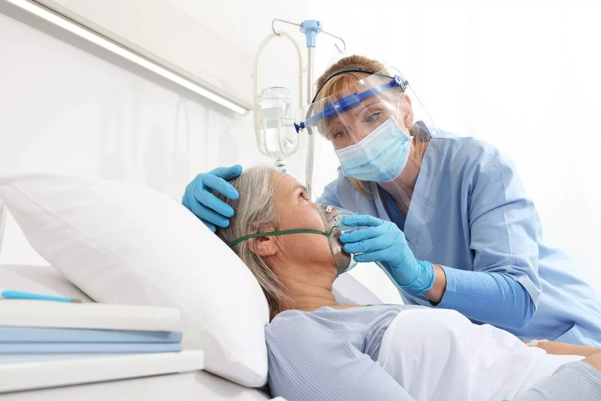 An individual in medical attire, including a mask and face shield, is attending to a patient in a hospital bed. The patient is lying down and appears to be receiving respiratory assistance, with both individuals wearing gloves. The hospital room is well-lit and equipped with standard medical apparatus.
