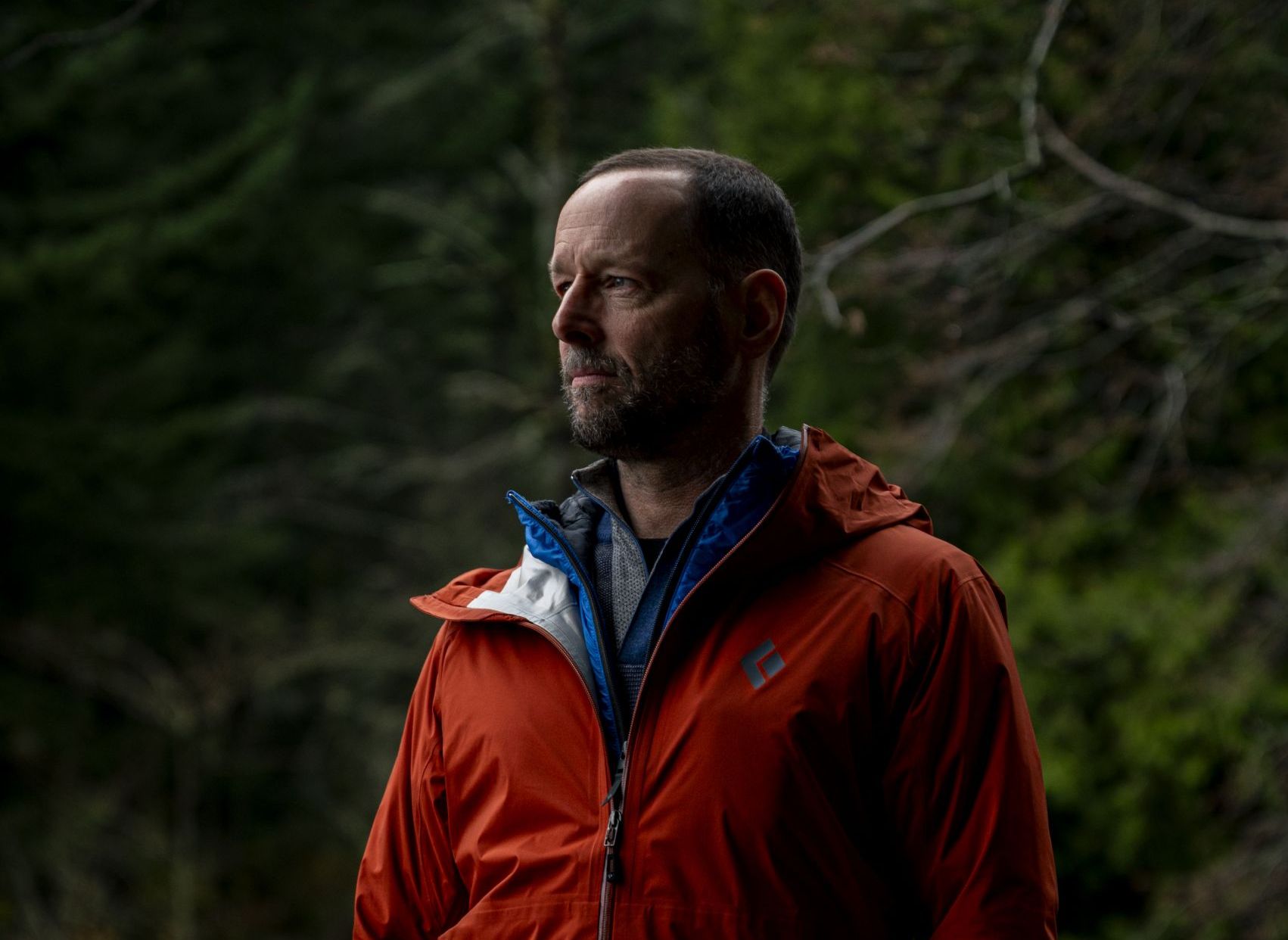 a portrait of Bill Donahue, against a treed background wearing a red rain coat