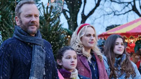 @SKYUK The production still shows 4 actors dressed in winter clothes with Christmas trees behind them. A red and yellow coloured fairground ride is on the right of the image. It is snowing.