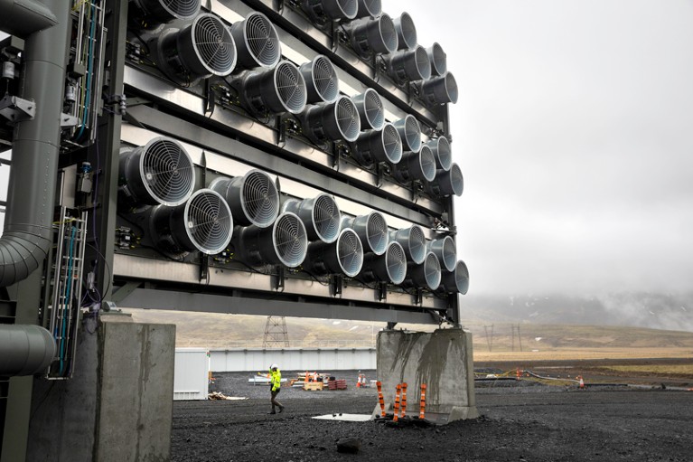 A worker in a hi-vis vest walks beneath a bank of fans many metres tall with circular fans arranged in three rows.