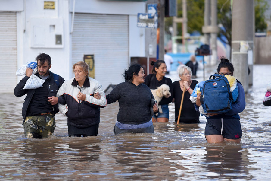 Photo of six people walking through thigh-deep brown water, carrying personal items or holding the arm of the person next to them, the day after a heavy storm in Bahia Blanca, 600 km south of Buenos Aires on March 8, 2025.