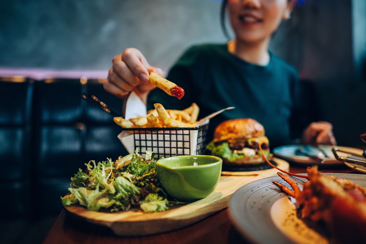 Cropped shot of young Asian woman eating freshly made delicious burger, dipping fries in ketchup on the dining table