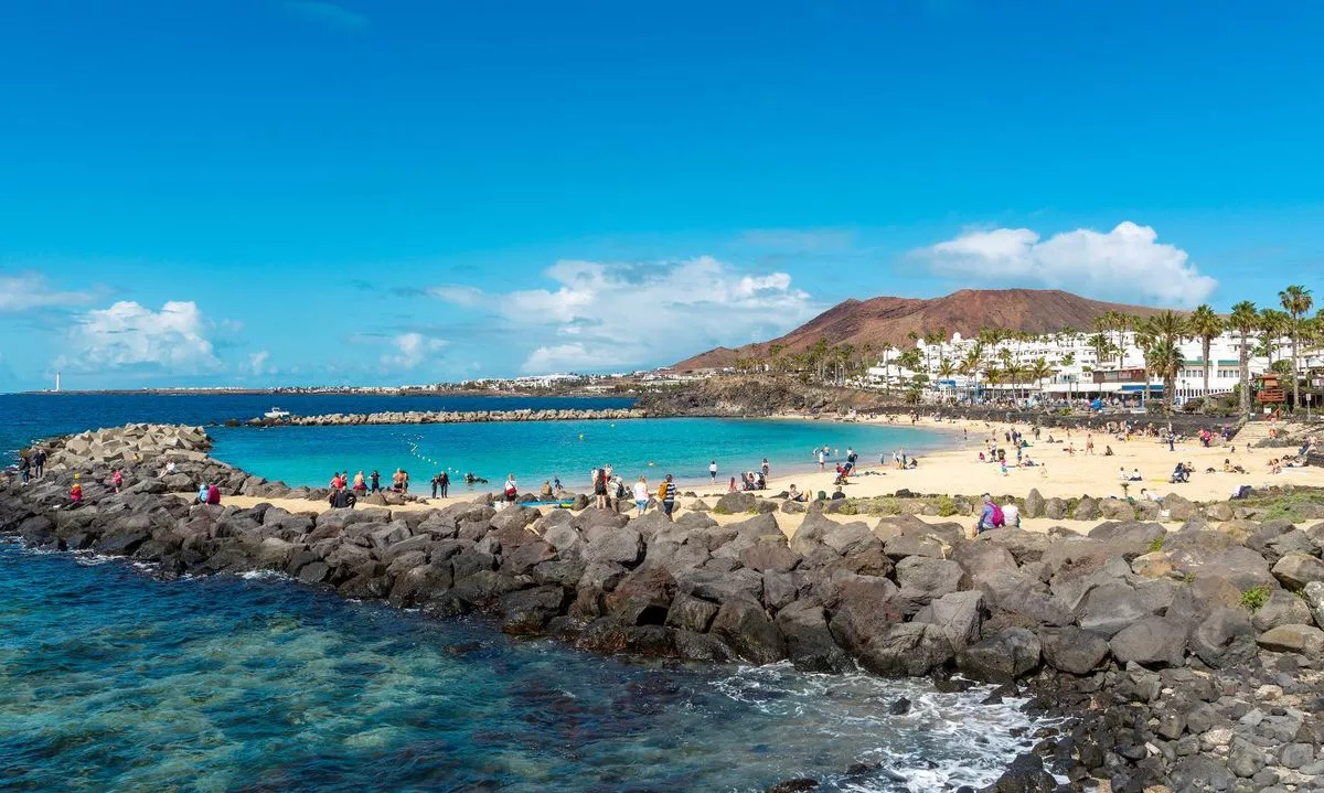 An aerial view of a coastal landscape featuring a serene beach with turquoise waters. The shoreline is bordered by rugged rocks, and in the distance, a hillside with several buildings is visible. The sky is clear with a few scattered clouds.