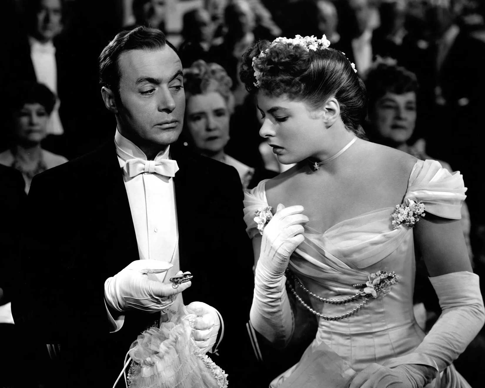 Charles Boyer, in a tuxedo with a white tie, holds a watch in his gloved hand and looks disapprovingly at Ingrid Bergman, looking distressed in a fancy dress and opera gloves in Gaslight