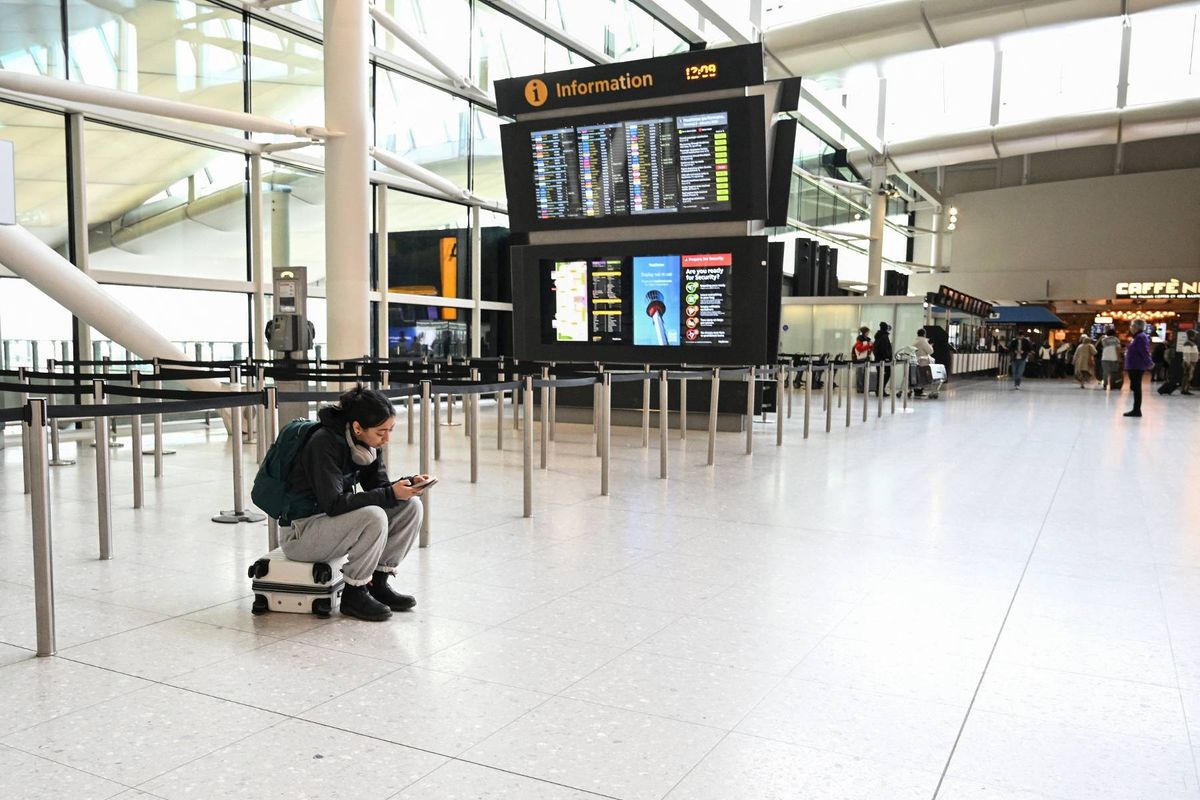 An individual is seated on the floor in a large, modern airport terminal, with a digital information board prominently displayed in the background. The terminal is largely empty, featuring clean lines and reflective surfaces.