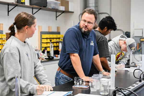 A teacher guides students during a hands-on science lab session, with equipment and materials spread across the lab bench.