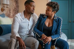 A man and a woman sitting on a bed, looking at each other intently.
