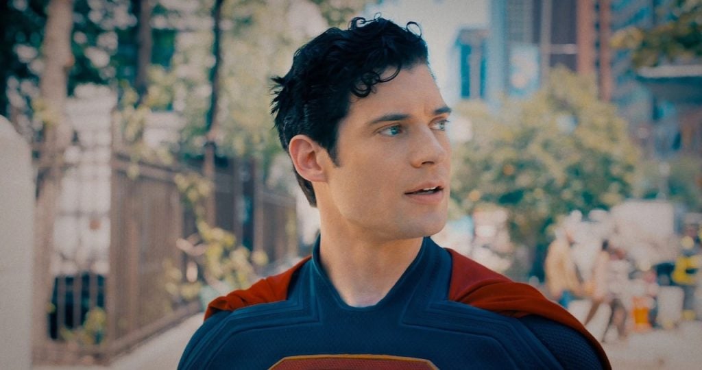 A man in a blue and red superhero costume with a red cape stands outdoors, looking to the side. Trees and buildings are visible in the background.