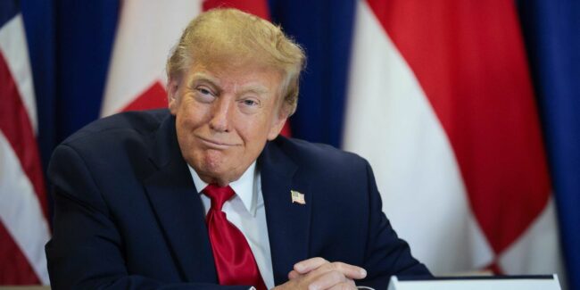 donald-trump-smiles-as-he-sits-at-a-table-with-a-sign-saying-united-states-on-it.jpg