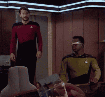 Two sci-fi characters in futuristic uniforms are in a starship setting; one stands, the other sits while wearing a high-tech visor