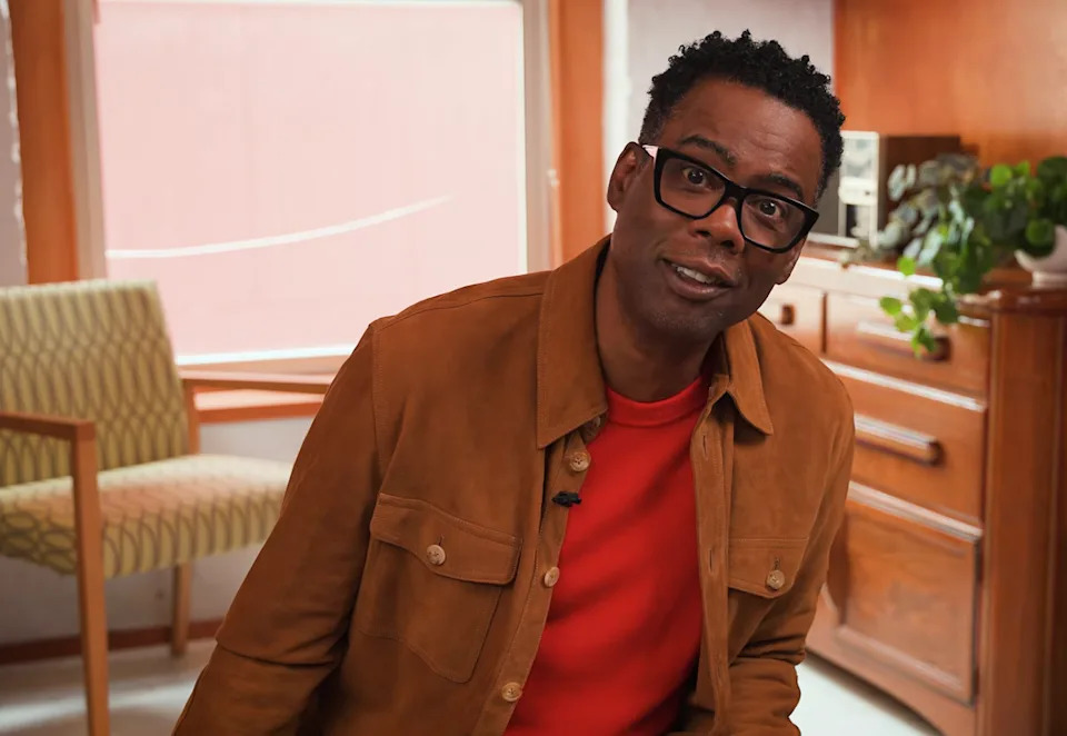 A person wearing glasses and a brown jacket over a red shirt sits in a cozy room with mid-century furniture