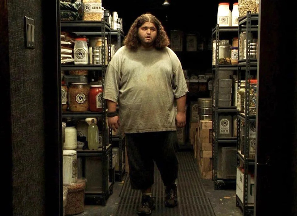 A person, Hurley, with long hair stands in a storage room filled with shelves of food and supplies, looking surprised or confused