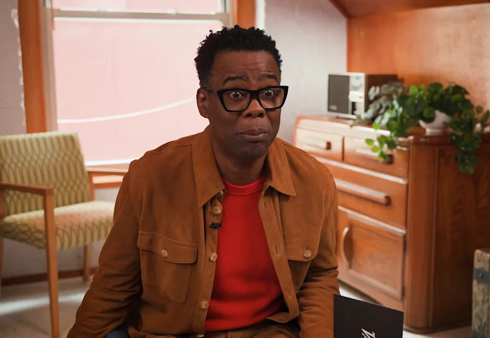 Person wearing glasses and casual clothing sits indoors, looking directly at the camera with a neutral expression