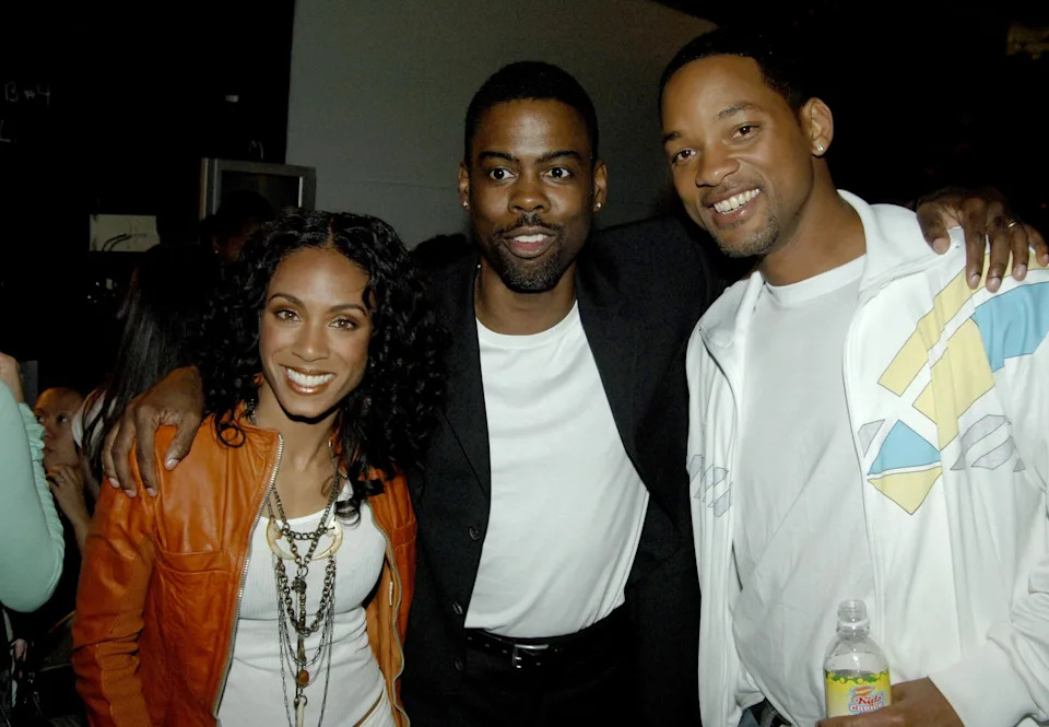 Three people posing indoors; the central figure is wearing a suit, and the others are dressed casually. They all appear happy