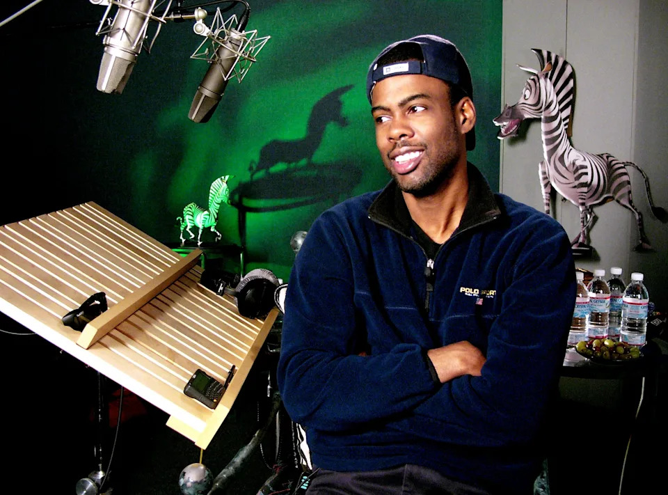 A person in a recording studio with microphones and zebra-themed decor, wearing a cap and a fleece jacket, appears to be speaking or listening