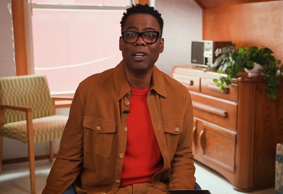 Person wearing glasses and casual jacket sits in a cozy room with wooden furniture and plants, speaking to the camera