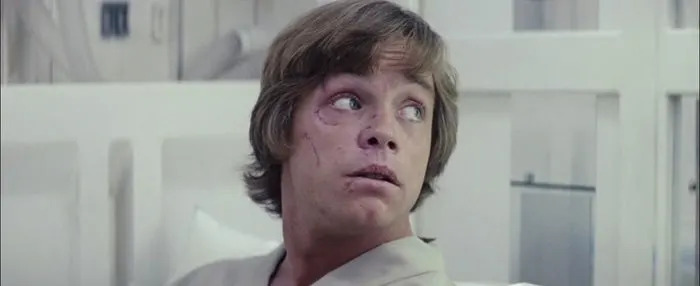 Luke, having short hair looking surprised while seated, wearing a light-colored robe, in a medical or sci-fi setting