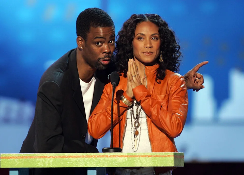 Two individuals stand at a podium, one wearing a blazer and the other in a leather jacket, speaking into microphones at an event