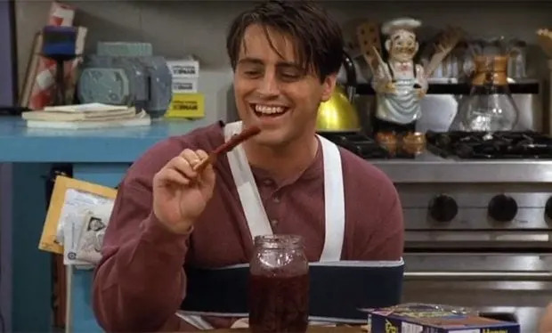 Joey with an arm in a sling happily dips a breadstick into a jar of red jam in a kitchen setting.
