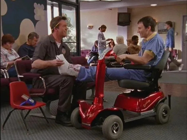 A man in a janitor uniform holds a foot cast of another man on a mobility scooter in a waiting room. They appear to be having a conversation