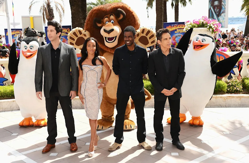 Group of four adults dressed in suits and formal wear, posing outdoors with penguin and lion characters from an animated movie