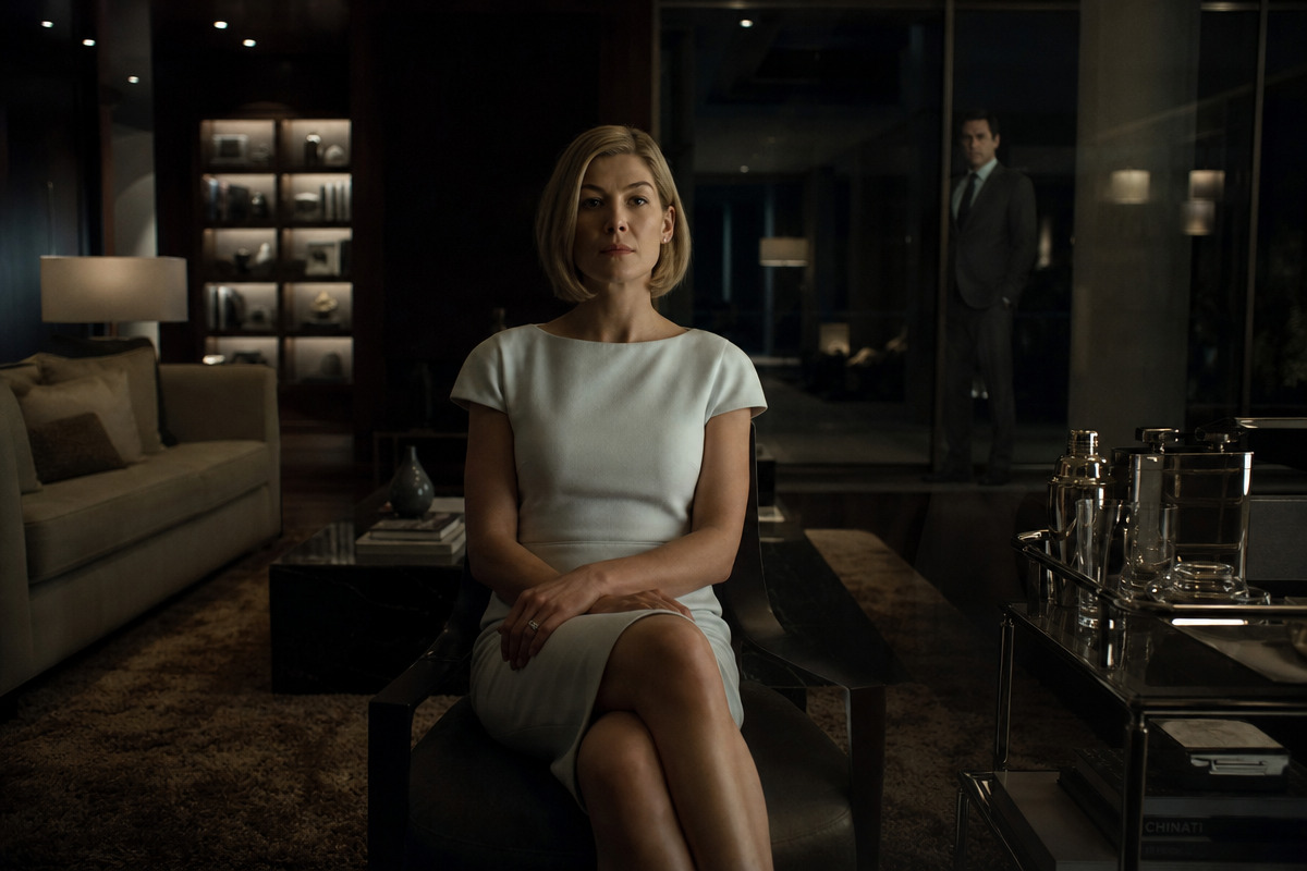 A poised blonde woman sits in a shadowy modern living room as a suited man appears reflected in distant glass.