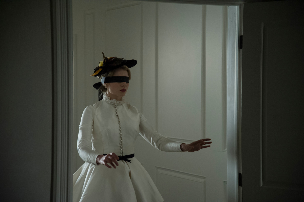 A woman in a long white dress and hat is blindfolded, carefully walking with outstretched arms inside a dimly lit, white-paneled room through an open doorway.