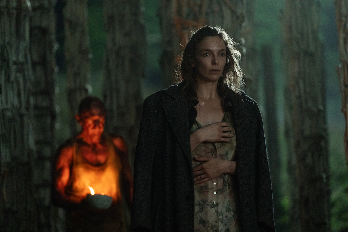 A woman in a coat stands in a dark, eerie forest, looking serious. Behind her, a person holding a lit candle adds a mysterious, tense atmosphere to the misty, wooded surroundings.