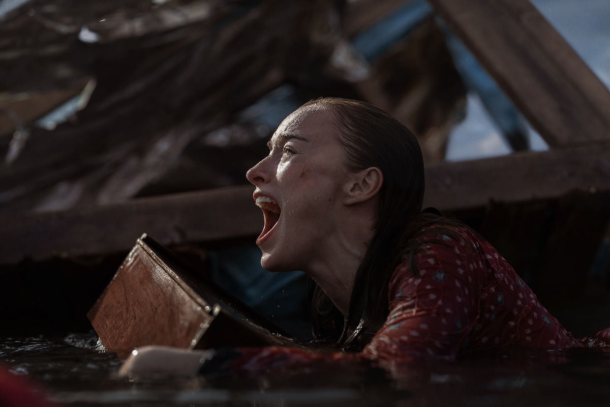 A woman in a red patterned shirt is in water, shouting desperately while holding onto debris, surrounded by wreckage, under a cloudy sky.