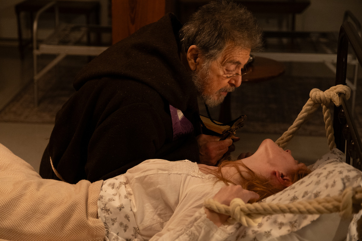 An elderly man in dark robes leans over a young woman who is tied to a bed, holding a crucifix. The scene is dimly lit, suggesting a dramatic or religious context, possibly an exorcism, set in a simple, sparsely furnished room.
