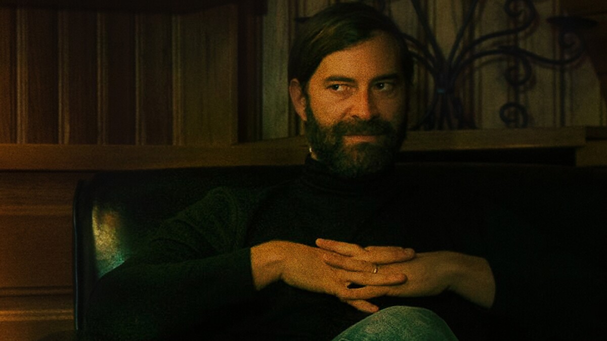 Bearded man with dark hair sits on a black leather chair in a warmly lit wooden room, wearing a black turtleneck and jeans, hands folded, looking to the side with a thoughtful or mischievous expression.