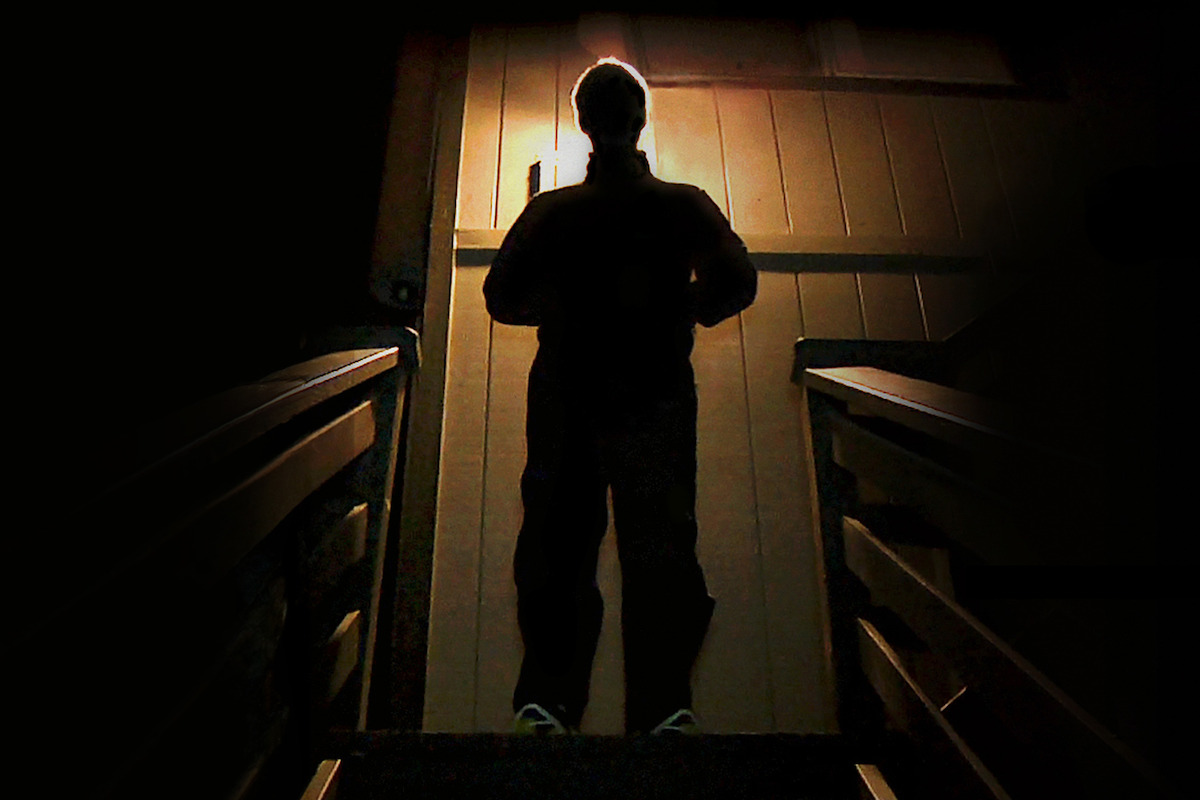 Silhouette of a person standing on stairs at night facing a closed door, illuminated from above by a single light, creating a mysterious and dramatic atmosphere.