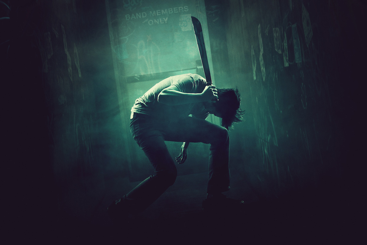 A person crouching in a dark, smoky hallway holding a machete, dramatic lighting casting shadows, posters on the walls, and a lit doorway labeled 