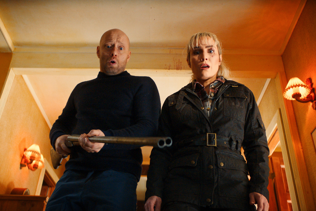 Two people stand indoors in a warmly lit hallway, looking down with surprised expressions. One person is holding a double-barreled shotgun, and both appear tense and alert, suggesting a dramatic or suspenseful moment.
