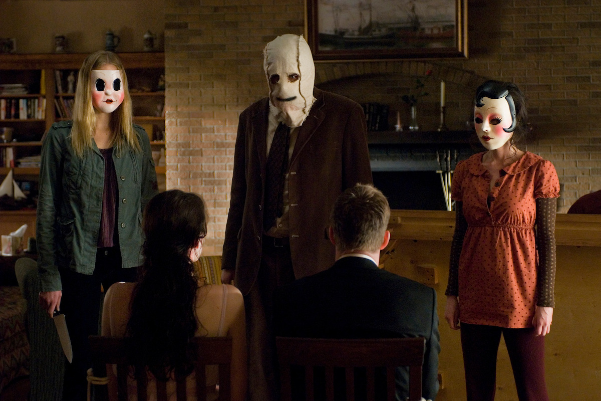 Three masked figures stand threateningly in a dimly lit living room facing two seated people with their backs to the camera, creating a tense and ominous atmosphere.