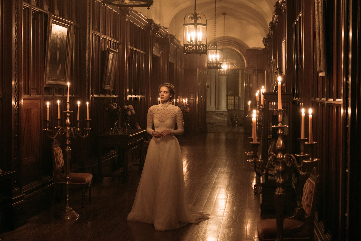 A woman in a vintage white gown stands in a grand, dimly lit hallway with wooden paneling, lit by candlelight and chandeliers, giving an elegant and mysterious atmosphere.