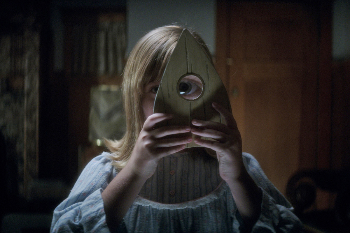 A young girl in a nightgown holds a wooden Ouija board planchette up to her eye in a dimly lit, old-fashioned room with wooden furniture and soft lighting.