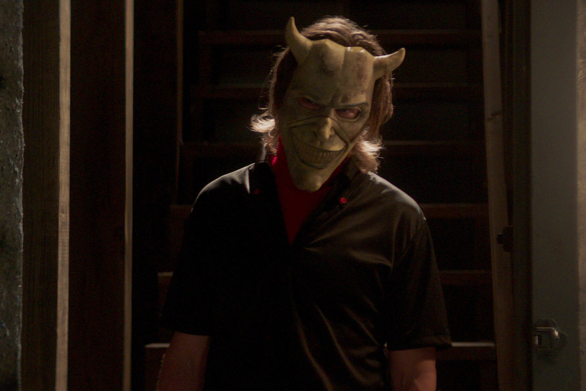 A person wearing a creepy horned mask stands in a dimly lit stairwell, with shadowy wooden steps behind, creating a suspenseful and eerie atmosphere.
