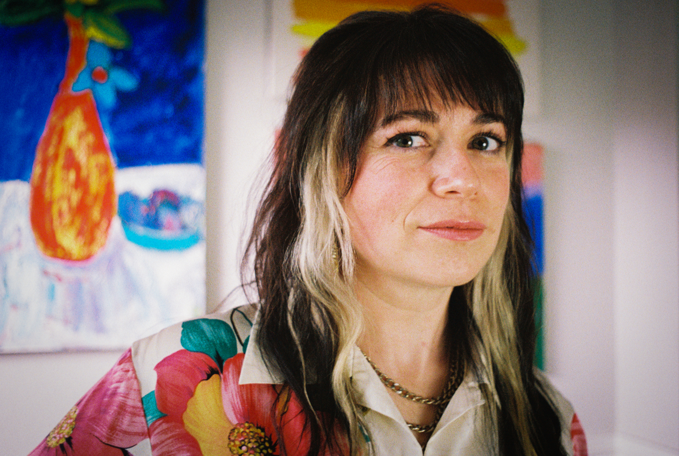 Sophie Shrand wearing a floral blouse looking into the camera while standing in front of brightly colored paintings.