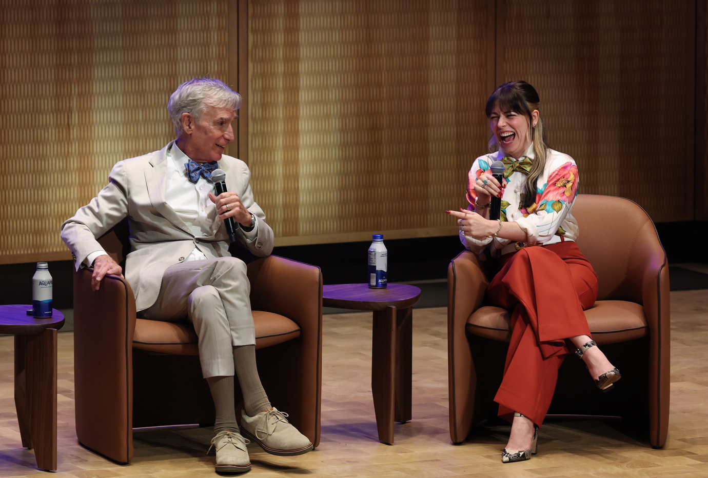 Bill Nye and Sophie Shrand sit in armchairs on a stage, laughing and holding microphones during an on stage conversation. They are both wearing bow ties.
