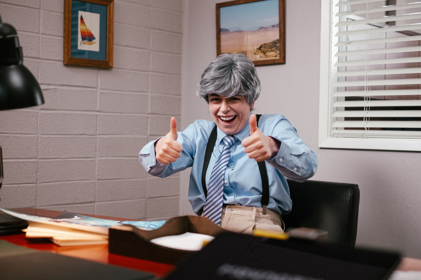 Sophie Shrand wearing a gray wig, a striped tie, suspenders and a blue button down grins and gives tow thumbs up from behind a desk in an office.
