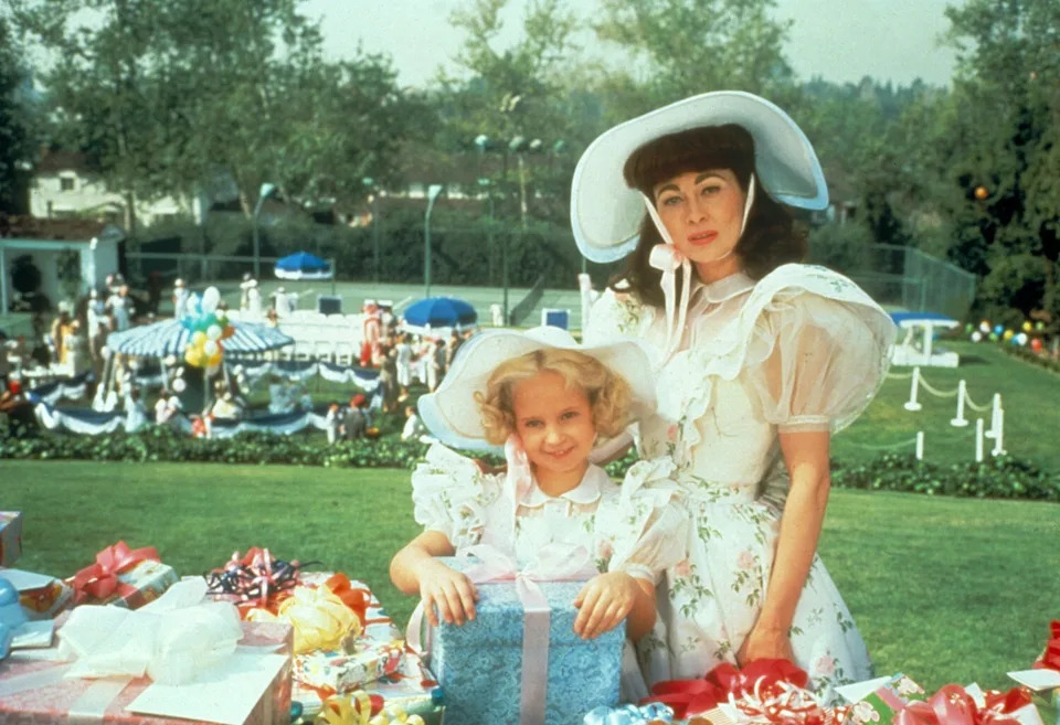 Faye Dunaway in a white hat as Joan Crawford stands behind child actress Mara Hobel at a lavish birthday party in Mommie Dearest