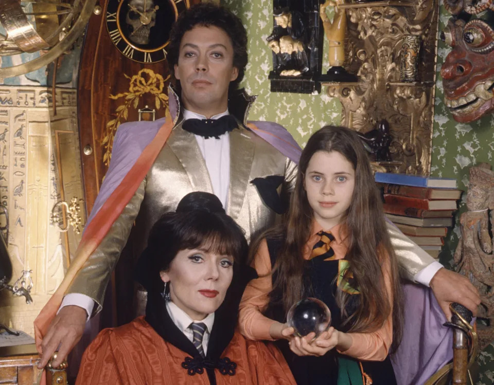 Tim Curry in a gold jacket and rainbow cape stands behind Diana Rigg and a young Fairuza Balk holding a crystal ball in The Worst Witch